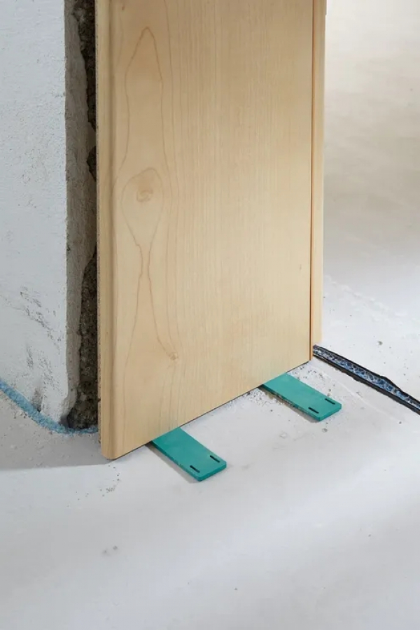 Detailný záber na zelené vyrovnávacie podložky zo sady Wolfcraft Pro umiestnené pod dvernou zárubňou pre presné nastavenie výšky od stonetrade.